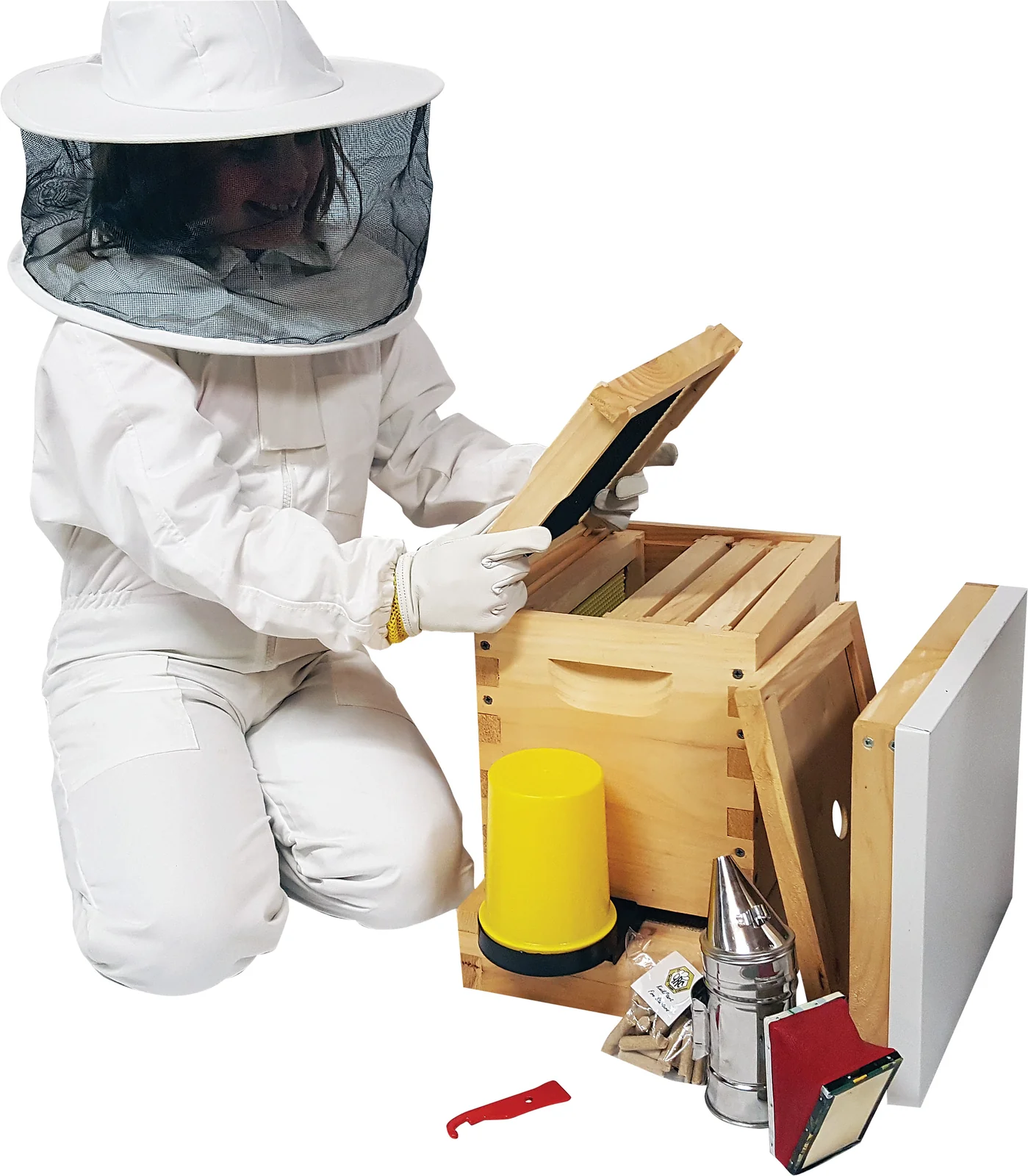 Person in a white bee suit and veil kneeling beside a wooden hive, with a smoker and hive tool