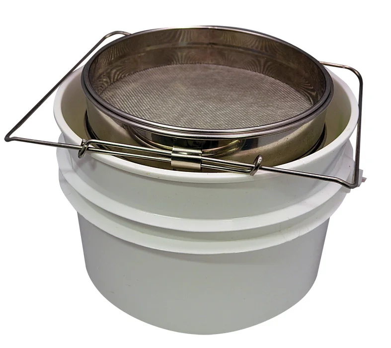 Double-mesh metal strainer resting over a white food-grade bucket