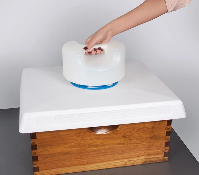 A hand resting on a white plastic feeder that sits on top of a wooden hive box